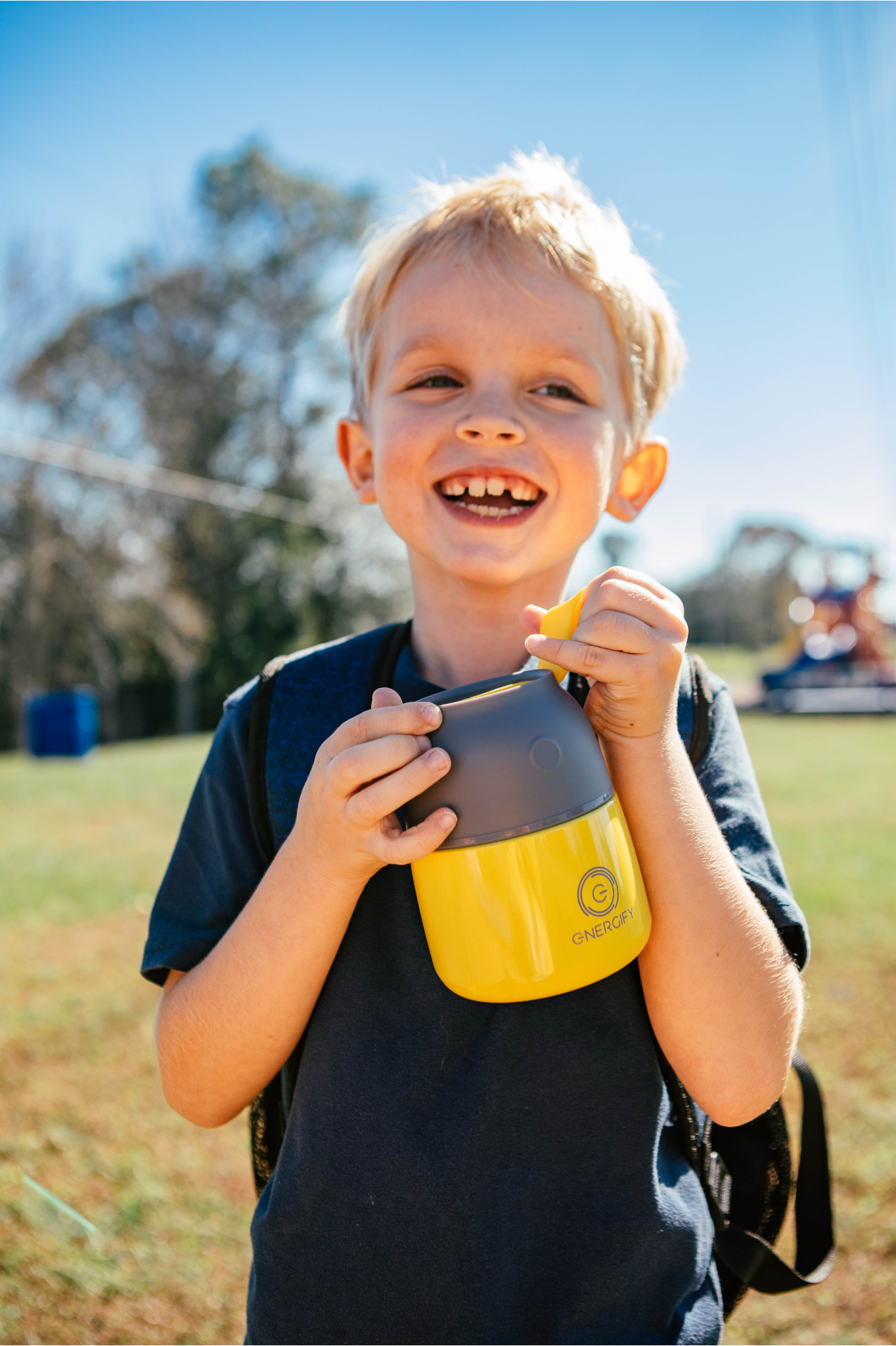 A young boy, approximately 8 years old, with short blonde hair and a wide, toothy grin proudly holds a 12 oz Starfruit Yellow Energify vacuum insulated food jar. He's wearing a dark blue t-shirt and a backpack with black and blue straps. The boy's two central incisors are not yet fully grown, he doesn't have lateral incisors, and he's looking to the side with a joyful expression, suggesting he's excited about the food or drink inside the jar.