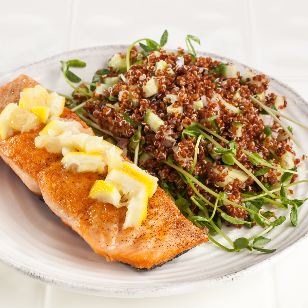 A plate of pan-seared salmon with lemon wedges on top, served with a side salad of quinoa, cucumber, and pea shoots.