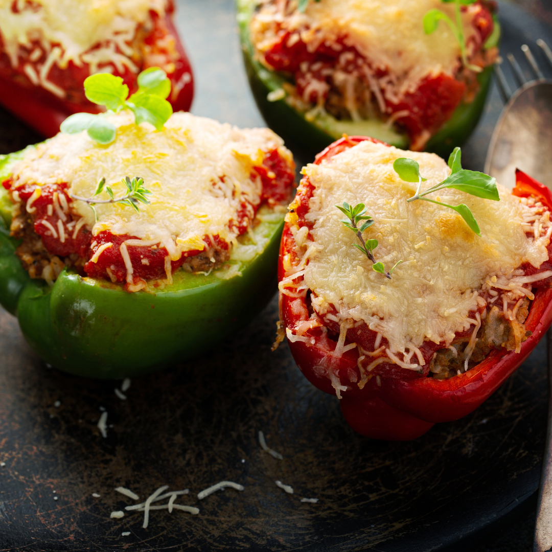 Stuffed bell peppers with a tomato-based filling, topped with melted cheese and fresh herbs. The peppers are a mix of red and green varieties, and are served on a dark plate.