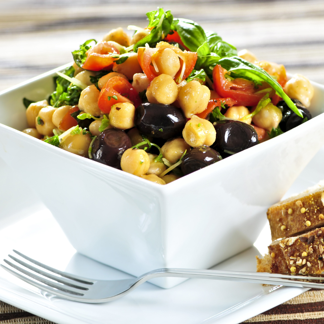 A bowl of chickpea salad with chopped tomatoes, olives, and fresh basil. The salad is served in a white square bowl with a fork and a slice of bread on the side.