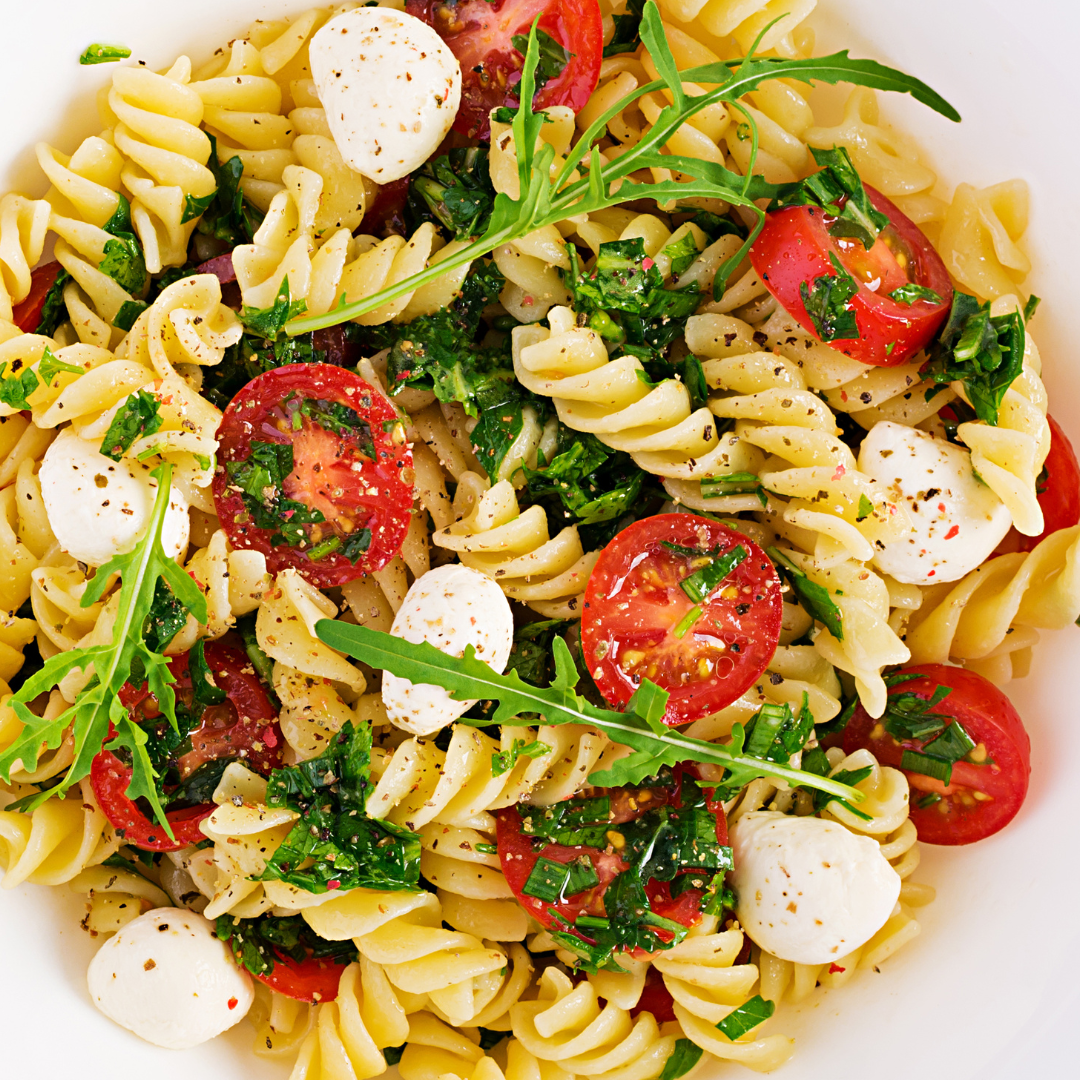 A bowl of pasta salad with fusilli pasta, cherry tomatoes, fresh mozzarella, and arugula. The salad is tossed with a dressing and seasoned with salt and pepper.