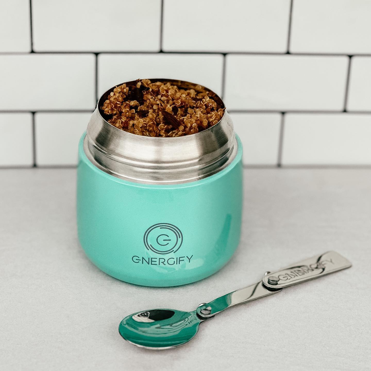12 oz mint green Energify vacuum insulated food jar with the lid open, revealing a portion of quinoa inside. A stainless steel spoon is placed next to the jar. The jar is sitting on a countertop with white tiles in the background.
