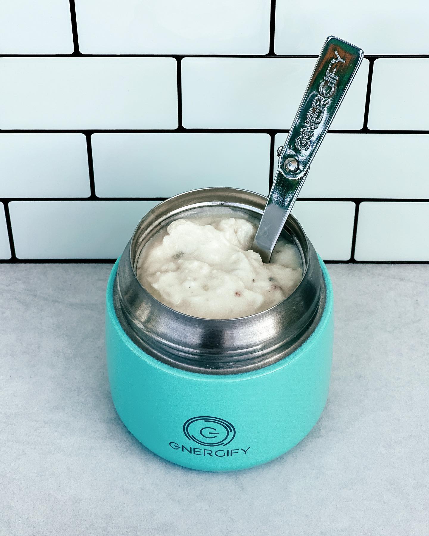 12 oz mint green Energify vacuum insulated food jar with the lid open, revealing a potato soup inside. A spoon is resting on the top of the mixture. The jar is sitting on a countertop with white tiles in the background.