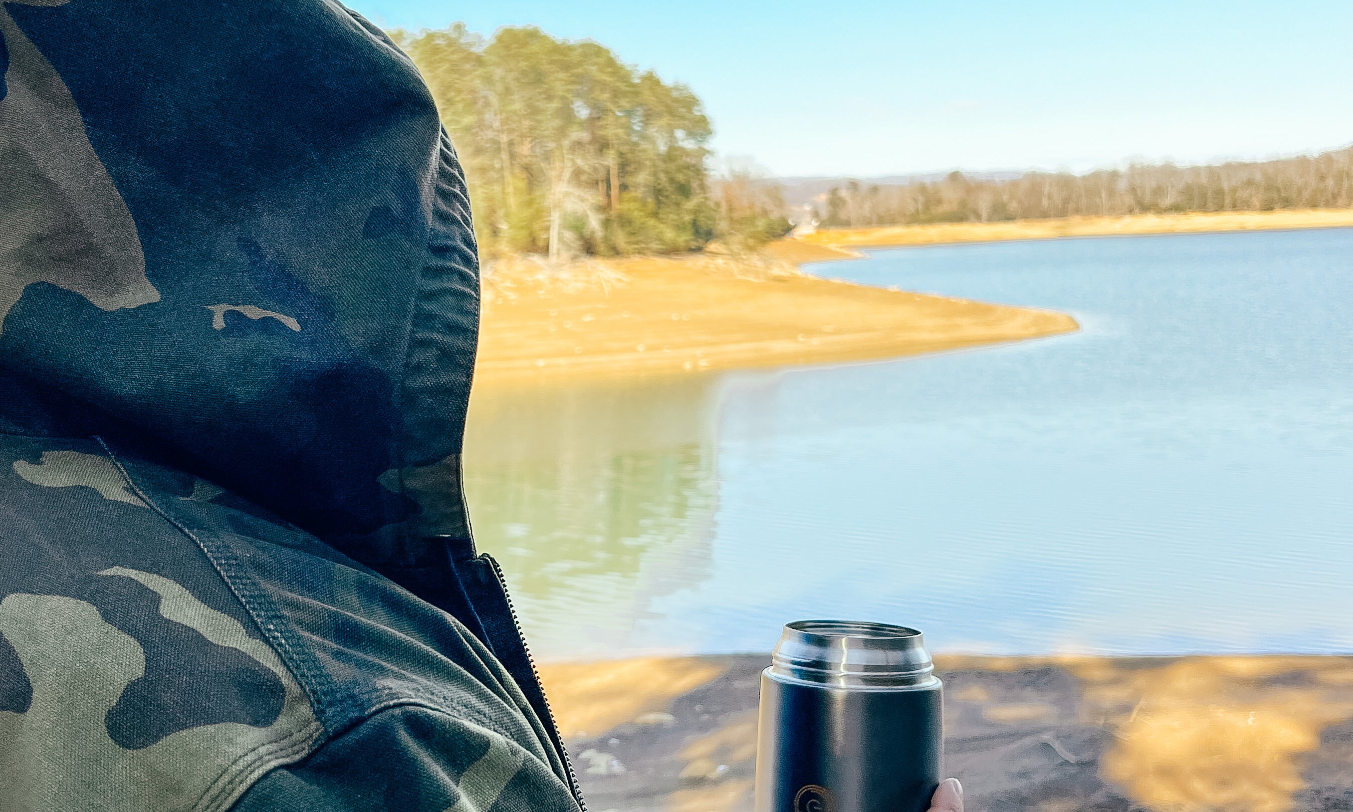 A man in a camo jacket and hood  on his head is holding in his right hand 24 oz Energify vacuum insulated food jar in Gray color. 