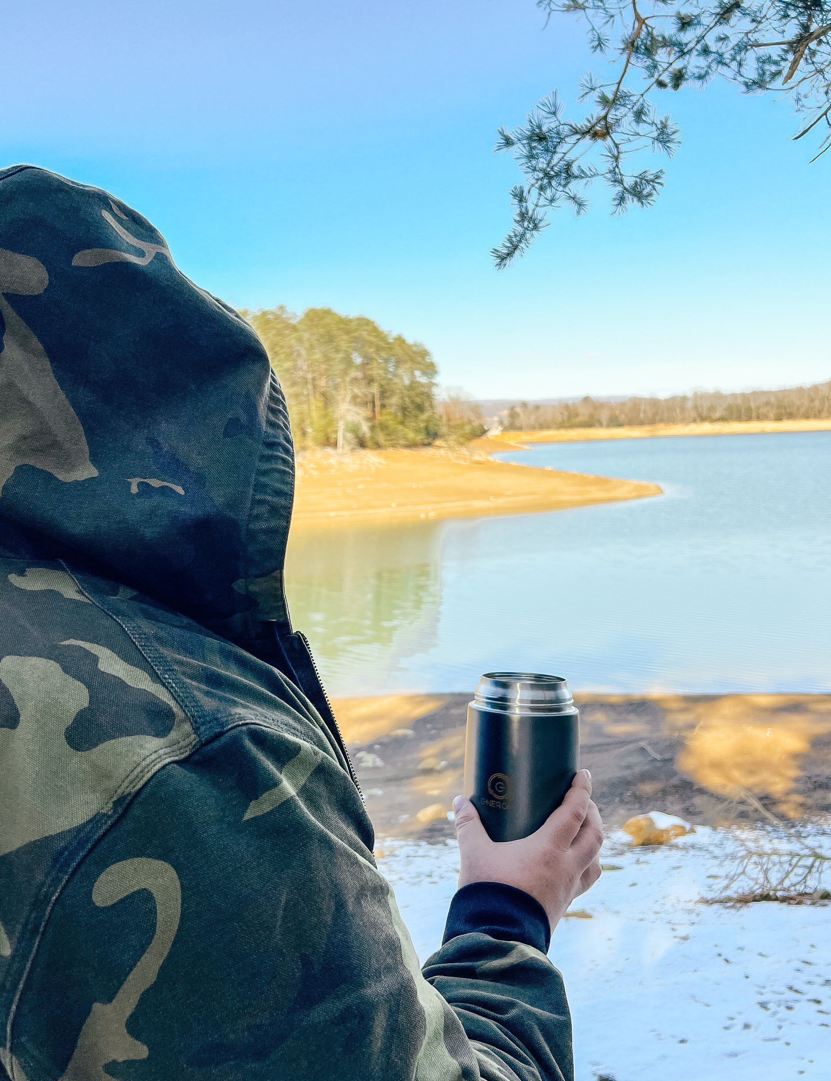 A man in a camo jacket and hood  on his head is holding in his right hand 24 oz Energify vacuum insulated food jar in Gray color. 