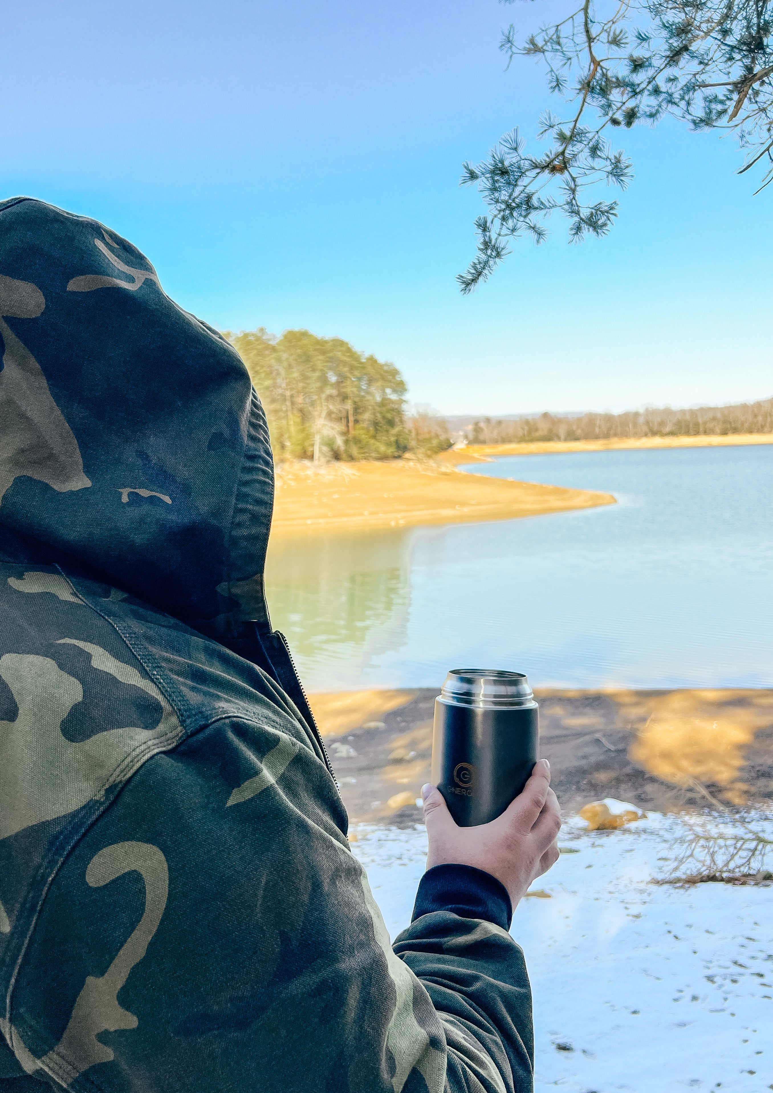 A man in a camo jacket and hood  on his head is holding in his right hand 24 oz Energify vacuum insulated food jar in Gray color. 