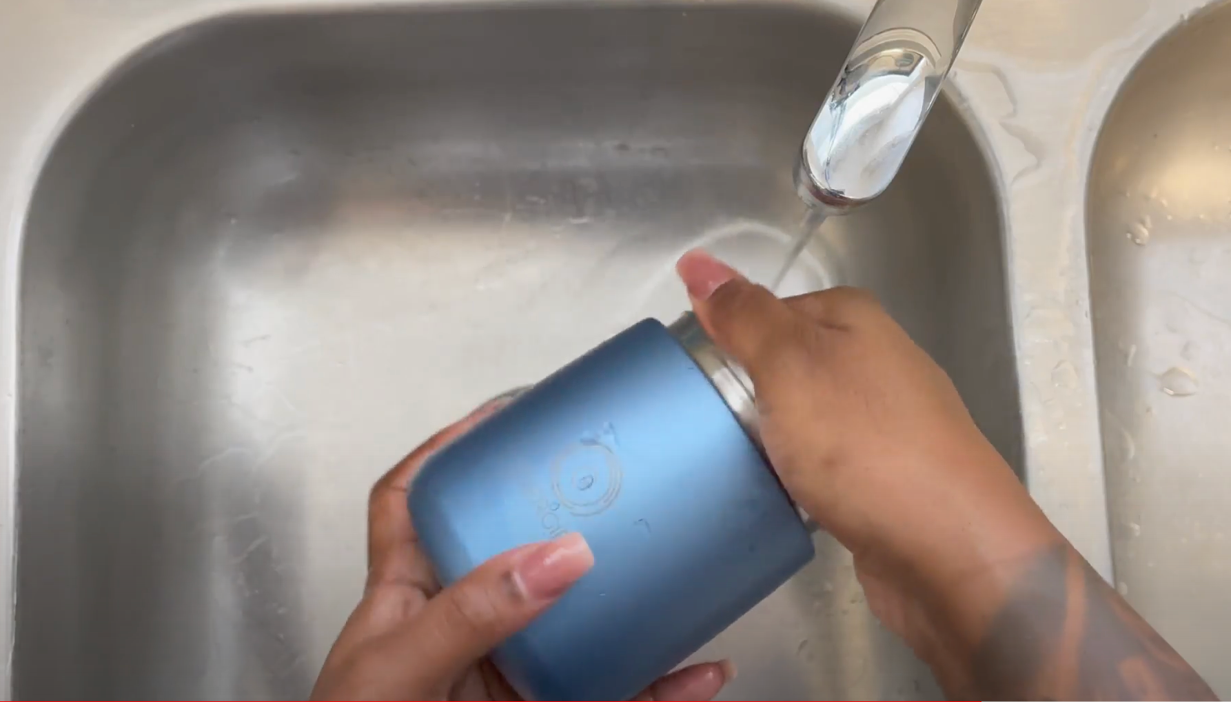 A person's hands washing a Shiny blue thermos in a kitchen sink. Water is running from the faucet and the jar is covered in soapy water.