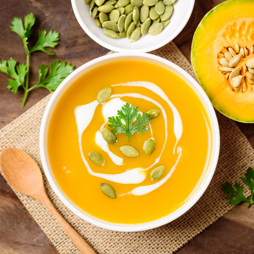 A bowl of pumpkin soup garnished with a dollop of cream, pumpkin seeds, and fresh cilantro. A half of a pumpkin and a bowl of pumpkin seeds are visible in the background.