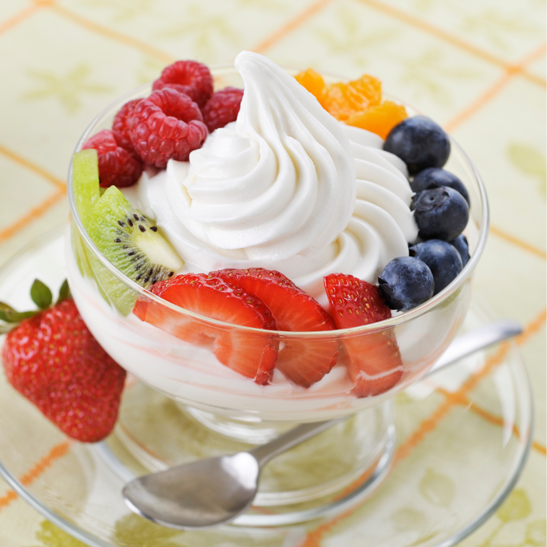 A bowl of frozen yogurt topped with a swirl of whipped cream and fresh fruit, including strawberries, raspberries, blueberries, and kiwi slices.