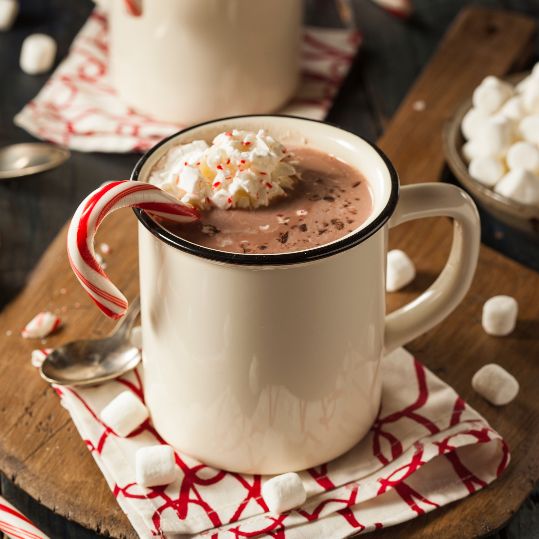 A mug of hot chocolate topped with whipped cream, crushed peppermint candy, and a candy cane. Marshmallows are scattered around the mug on a wooden board.